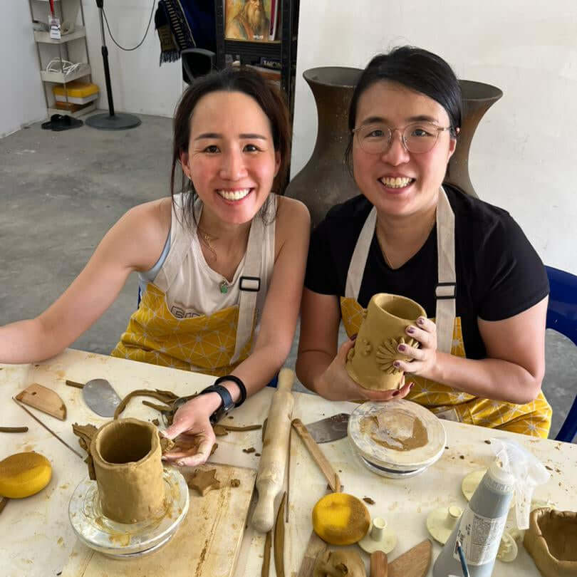 Two women smiling in pottery studio, enjoying their pottery studio membership while creating clay mugs.